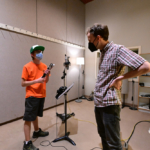 A student holding a folded-up cane and wearing a face mask stands next to a microphone, talking with Daniel, also wearing a mask.