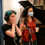 A student wearing headphones and standing in front of a microphone covers their mouth as if laughing. Jessica stands behind, wearing a face mask and holding a paper cup.