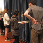 Three people wearing headphones standing in a row, microphones visible in front of two of them. The closest has his hands on a music stand with a piece of paper on it. Daniel Guenther stands on the right, facing them.