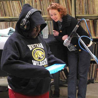 A student wearing headphones holds an ice try in front of a microphone. Christine Brown looks on, smiling.