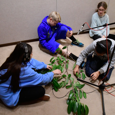 Students sit on studio floor holding leaves and branches near a microphone.