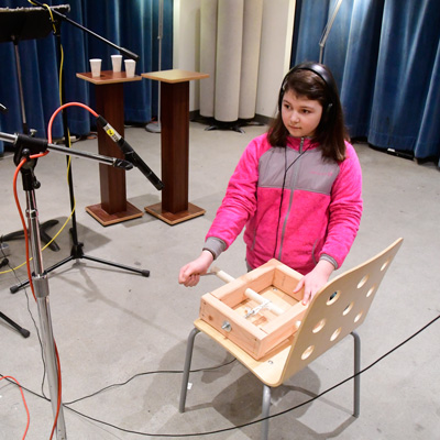 A student wearing headphones kneels, turning a handle on a wooden device sitting on a wooden chair. A microphone points toward the device.