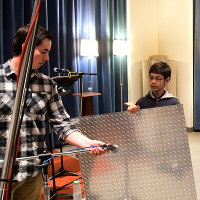 A student holds a large sheet of corrugated steel; engineer adjusts a microphone next to it.