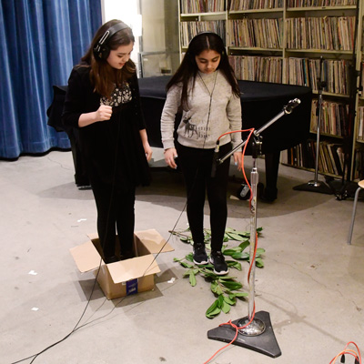 Two students in a studio wearing headphones, one standing in a box, the other on leafy branches. A microphone points toward the floor.