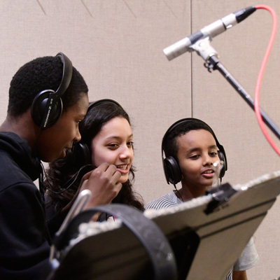 Three students wearing headphones stand together, a microphone overhead.