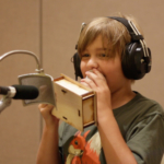 A student at a microphone, holding a wooden box up to their mouth and wearing headphones.