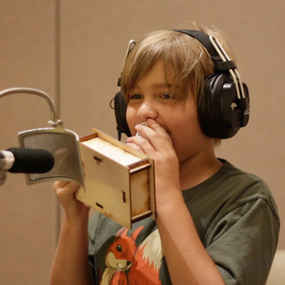 A student at a microphone, holding a wooden box up to their mouth and wearing headphones.