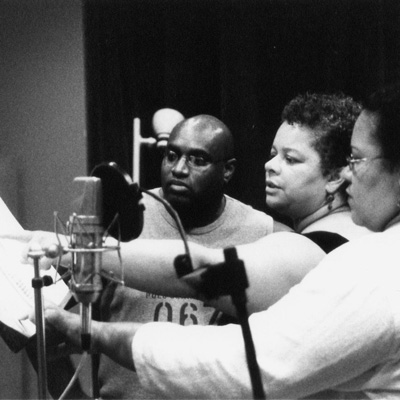 Black and white photo of three people standing a a microphone, one holding a page at arm's length while another points to the page.