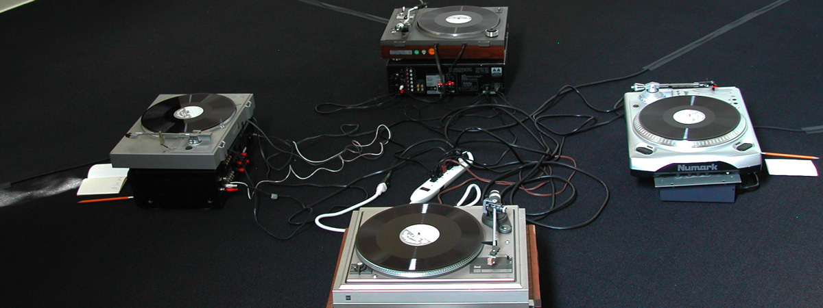 Four LP turntables arranged in a square on a black-carpeted floor, each with a record on it.