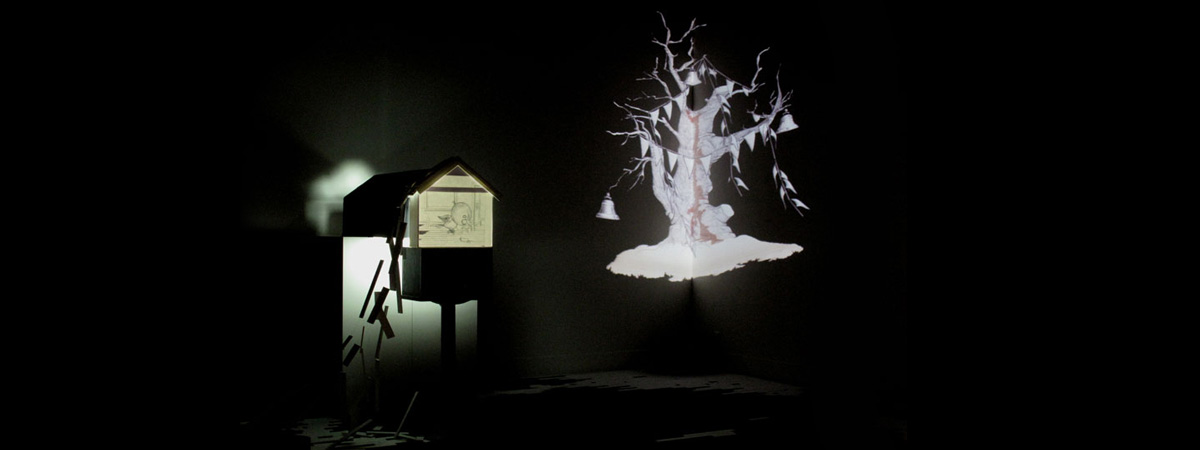 View of the installation So Long, a dark room with a paper sculpture showing a projected illustration of a bird, and a large projection of of a tree with bells and ribbon in the corner of the room.