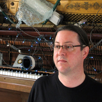 Photo of Tim Root, with an open upright piano and other musical equipment in the background.