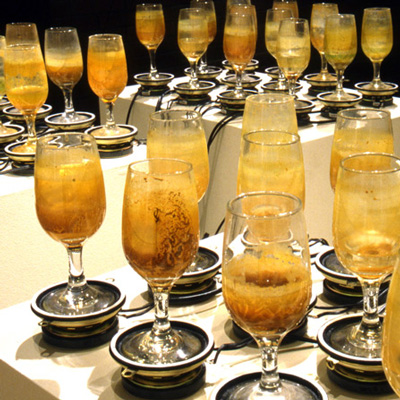An arrangement of wine glasses balanced on small speakers on top of pedestals, with murky orange liquid in them.