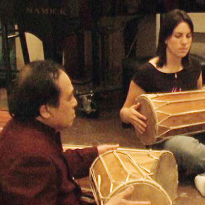 Photo of I Wayan Sinti, seated on the floor, playing a hand drum. On the right, a student also plays a hand drum.