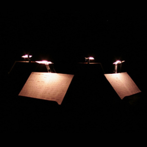 Four music stands in the dark, two facing the camera and two facing away, each lit from above.