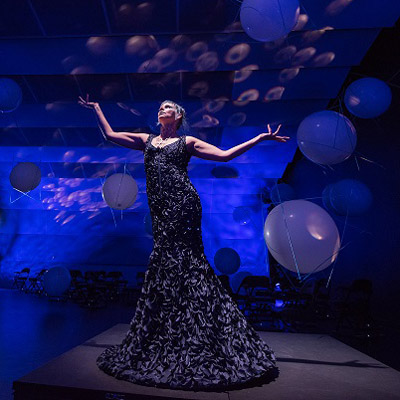 KT Niehoff in a long black gown on a platform in a blue-lit room with balloons behind her.
