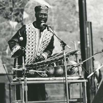 Black and white photo of a man playing West African percussion instruments on a stage.