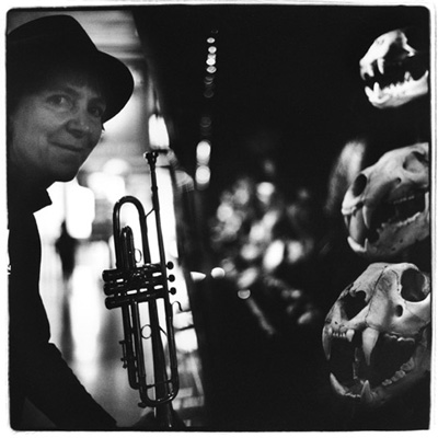 A black and white photo. Lesli Dalaba wearing a hat and holding a trumpet on the left. On the right, three animal skulls.