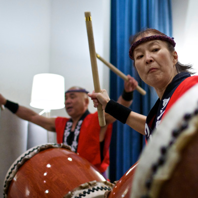 Two people wearing red and black clothing and black headbands playing large drums with large wooden sticks.