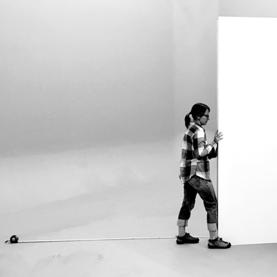 Black and white photo of Tania Kupczak adjusting a white wall, with an extended tape measure on the floor.