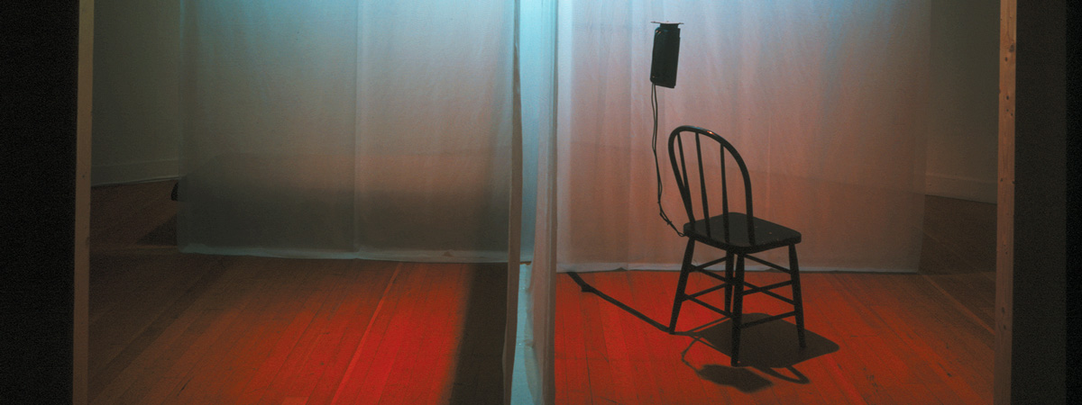 A room divided into four by a translucent fabric. In the foreground on the right, a black wooden chair attached to an electronic device in one of the quadrants.
