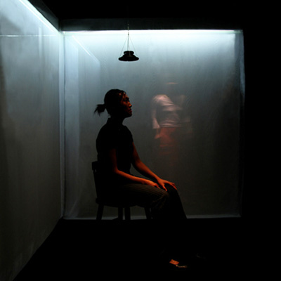 A person sits on a chair in a dark space, with an illuminated semi-transparent screen behind them. A small speaker hangs overhead.