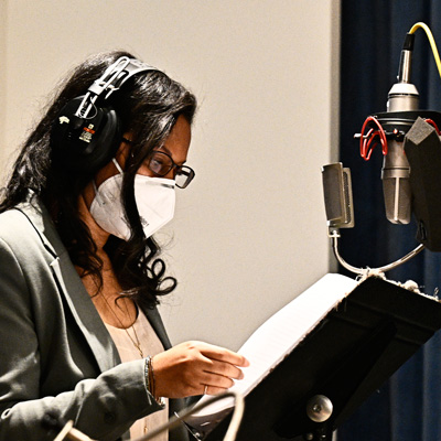 Danielle Hayden wearing headphones and a white facemask, standing at a microphone looking at a sheet of paper on a music stand.