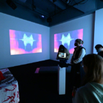 Several people standing in a dimly late space with projected images on the walls. One holds a laptop computer with someone visible on Zoom on the screen.