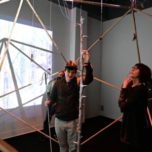 Sasha Petrenko and a student with a white cane stand inside a large, geometric structure made of wooden rods in a darkened room. An image is projected on the wall behind them.