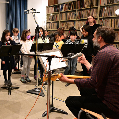 Tito Ramsey seated, holding a guitar. Children standing at music stands facing him. Microphones overhead and in front of Tito.