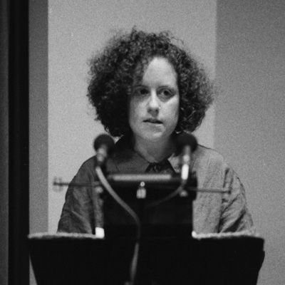 Black and white photo of Becka Mara McKay, standing at a lectern with two microphones.