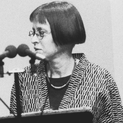 Black and white photo of Deborah Woodard, standing in front of two microphones and looking to the left.