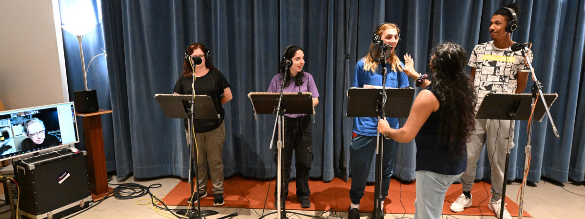 A group of four students stand at music stands and microphones as an engineer faces them, adjusting a microphone. A computer monitor on the left shows a face in an online meeting.
