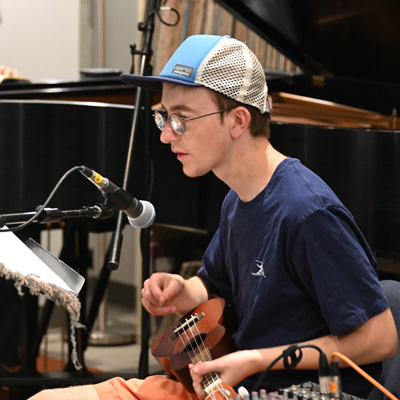 A student wearing glasses and a baseball cap sits holding an ukulele, a microphone in front of him.