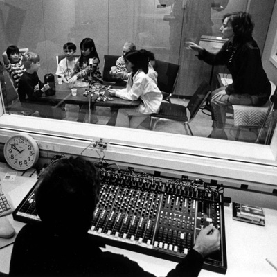 Black and white photo of students and an adult in a recording studio while someone at a mixing board looks on from the control room through a window.