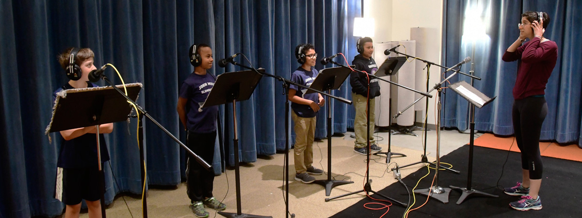 Four students wearing headphones at microphones with music stands in front of them, while a vocal coach stands on the right facing them.