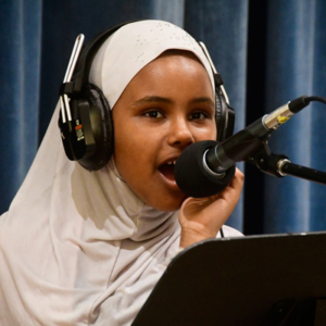 A student in a hijab wearing headphones leans into a microphone to speak.