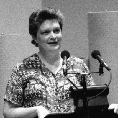 Black and white photo of Kathryn Owen standing at a lectern, two microphones in front of her.