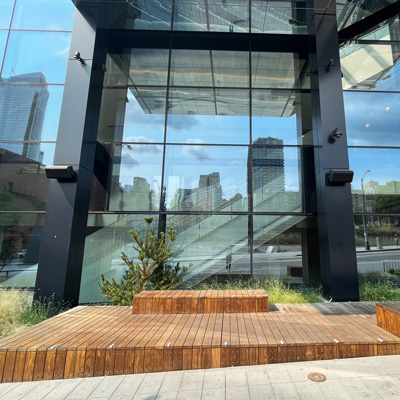 Wooden bench and platform adjacent to a glass and metal building in the background. Speakers mounted on black columns over the bench.