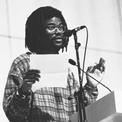 Black and white photo of Eben Eldridge, holding a sheet of paper and standing at a microphone.