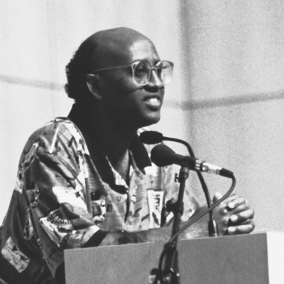 Black and white photo of Jamal Gabobe, standing at a lectern with two microphones in front of him.