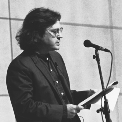 Black and white photo of Kimball MacKay-Brook, holding some pages and standing at a microphone.