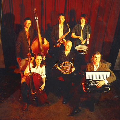 A group of musicians with upright bass, saxophone, drums, cello, french horn, and accordion, standing in front of a red curtain.