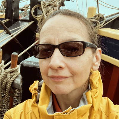 Joann Farias wearing sunglasses and a yellow raincoat, with a ship's rigging in the background.