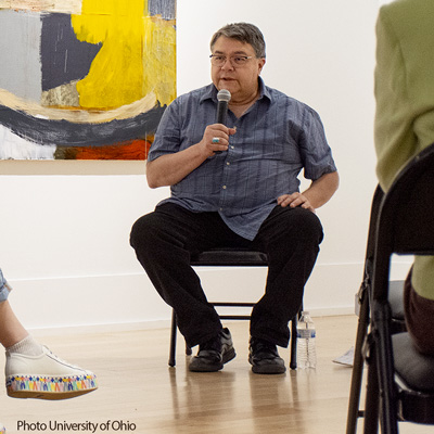 Photo of John Feodorov, seated and holding a microphone, with a colorful painting behind him.