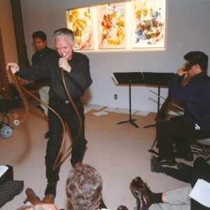 A gallery space with backlit paintings on a wall, people seated in the foreground. Stuart Dempster blows into a long, winding tube and Loren Dempster plays cello.
