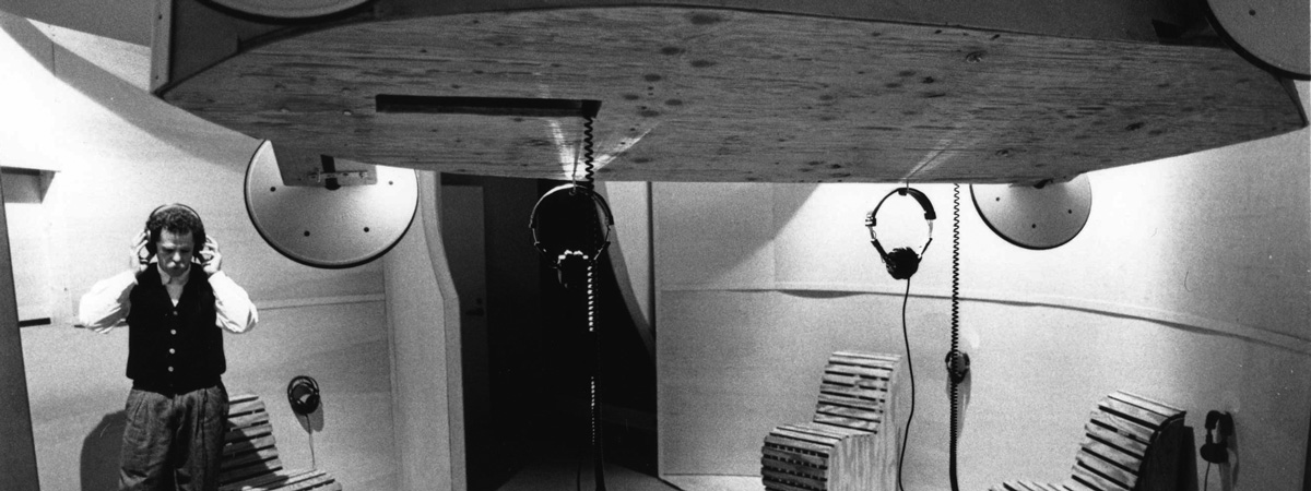 Black and white photo of Don Fels wearing headphones in a gallery space with a large plywood sculpture and several hanging pairs of headphones.