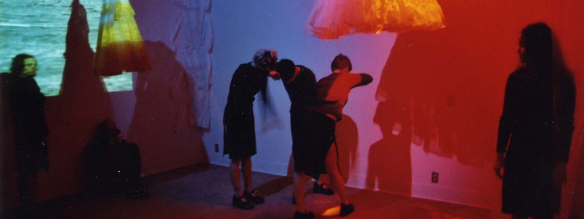 A red and purple lit gallery space with projections, sculptural objects, and dancers performing.