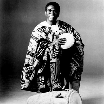 Obo Addy holding an African hand drum, with his foot on another drum lying on the floor.