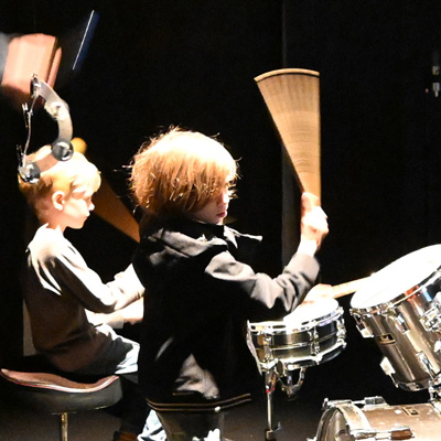 Two youth at a drum kit in a darkened room, one swinging a drum stick.