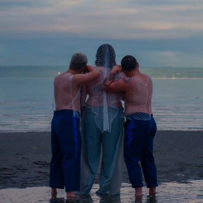 Three people looking out at the ocean from behind, leaning on each other.